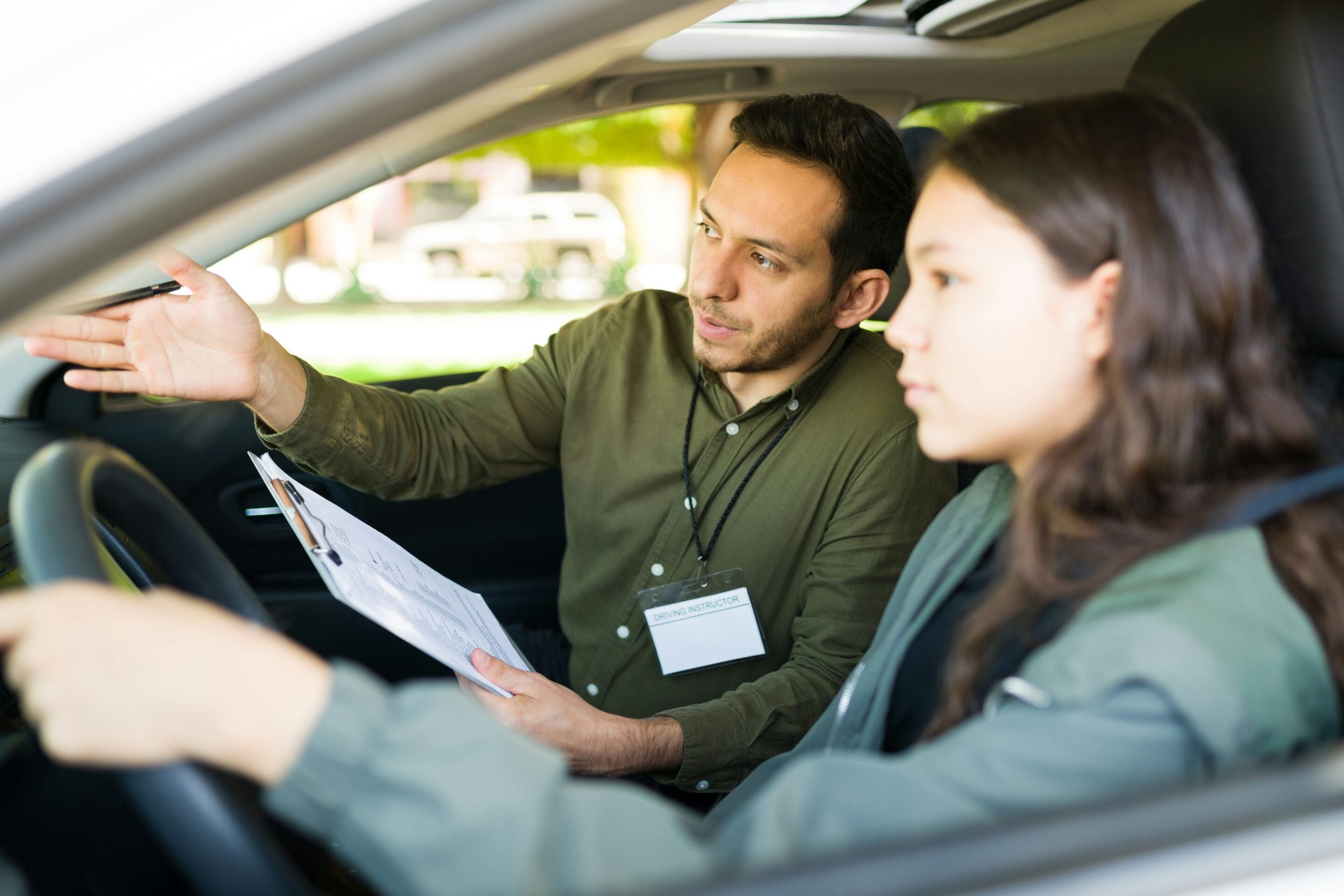 Ein Fahrlehrer sitzt auf dem Beifahrersitz, hält ein Klemmbrett in der Hand und zeigt nach vorne, während eine junge Frau auf dem Fahrersitz aufmerksam zuhört und das Lenkrad hält.