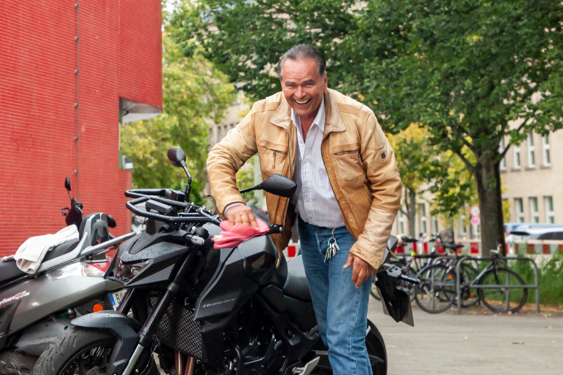 Ein lächelnder Mann in hellbrauner Jacke und Jeans wischt ein schwarzes Motorrad mit einem rosa Tuch ab, während er im Freien in der Nähe eines roten Gebäudes und von Bäumen steht. Im Hintergrund sind Fahrräder geparkt.