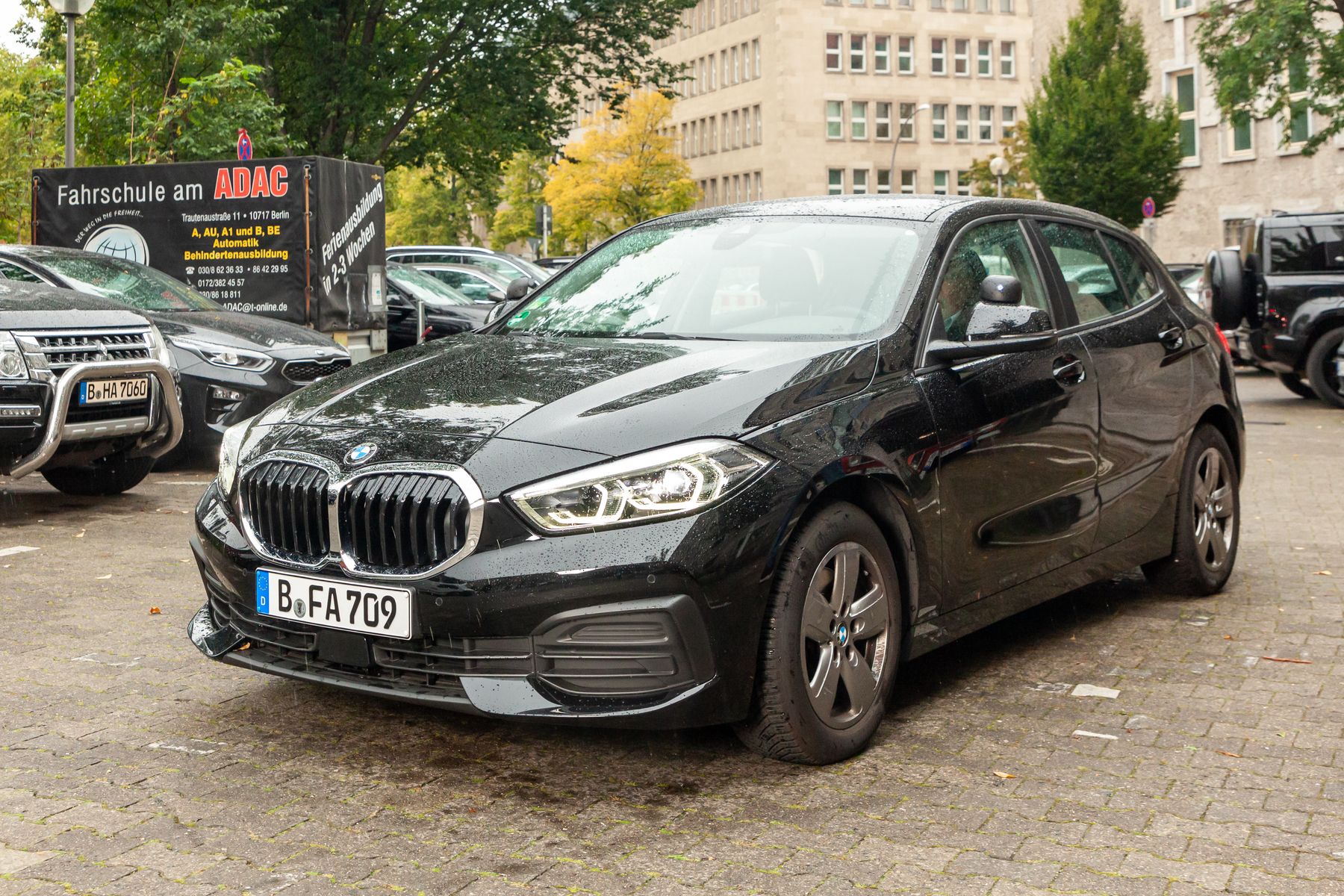 Ein schwarzer BMW mit Schrägheck ist auf einer Kopfsteinpflasterstraße vor einem Fahrschulschild geparkt, im Hintergrund sind andere Autos und Bäume zu sehen.
