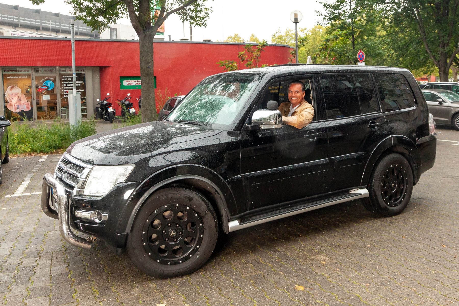 Ein lächelnder Mann, der auf dem Fahrersitz eines schwarzen Geländewagens sitzt, der auf einem gepflasterten Grundstück mit Bäumen und einem roten Gebäude im Hintergrund geparkt ist.