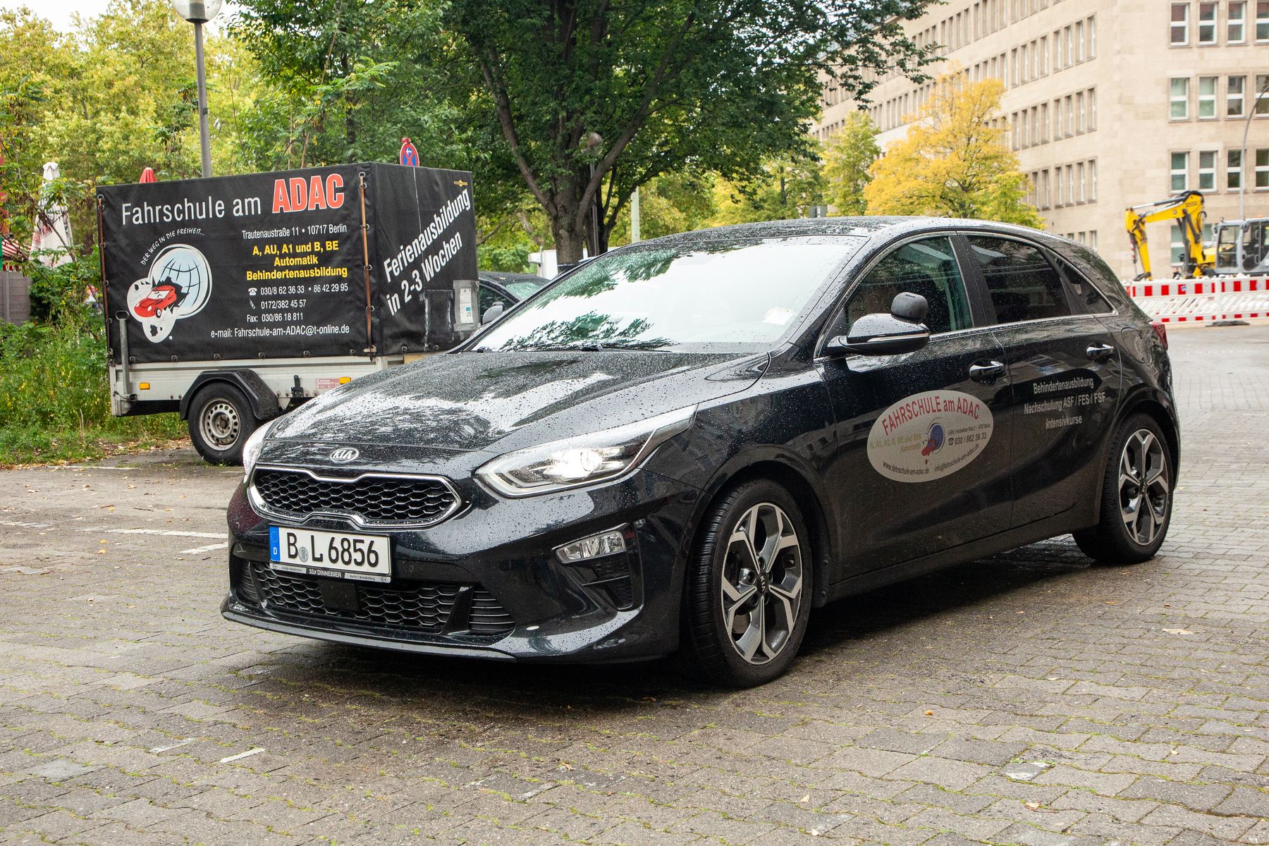 Ein schwarzer Kia mit Schrägheck und Fahrschullogo ist auf einer nassen Straße geparkt, im Hintergrund sind ein Fahrschulbus und städtische Gebäude zu sehen. Auch Bäume und Bauzäune sind zu sehen.
