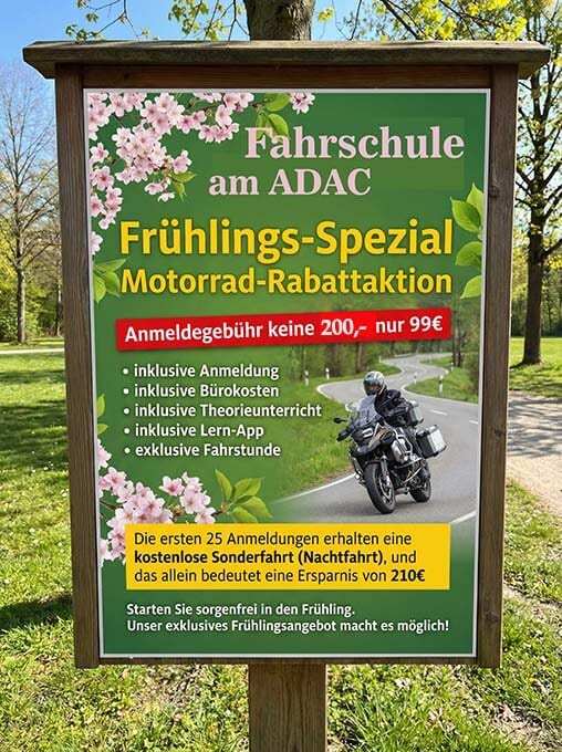 ein schild am straßenrand unter bäumen wirbt für eine frühjahrsaktion einer fahrschule: 