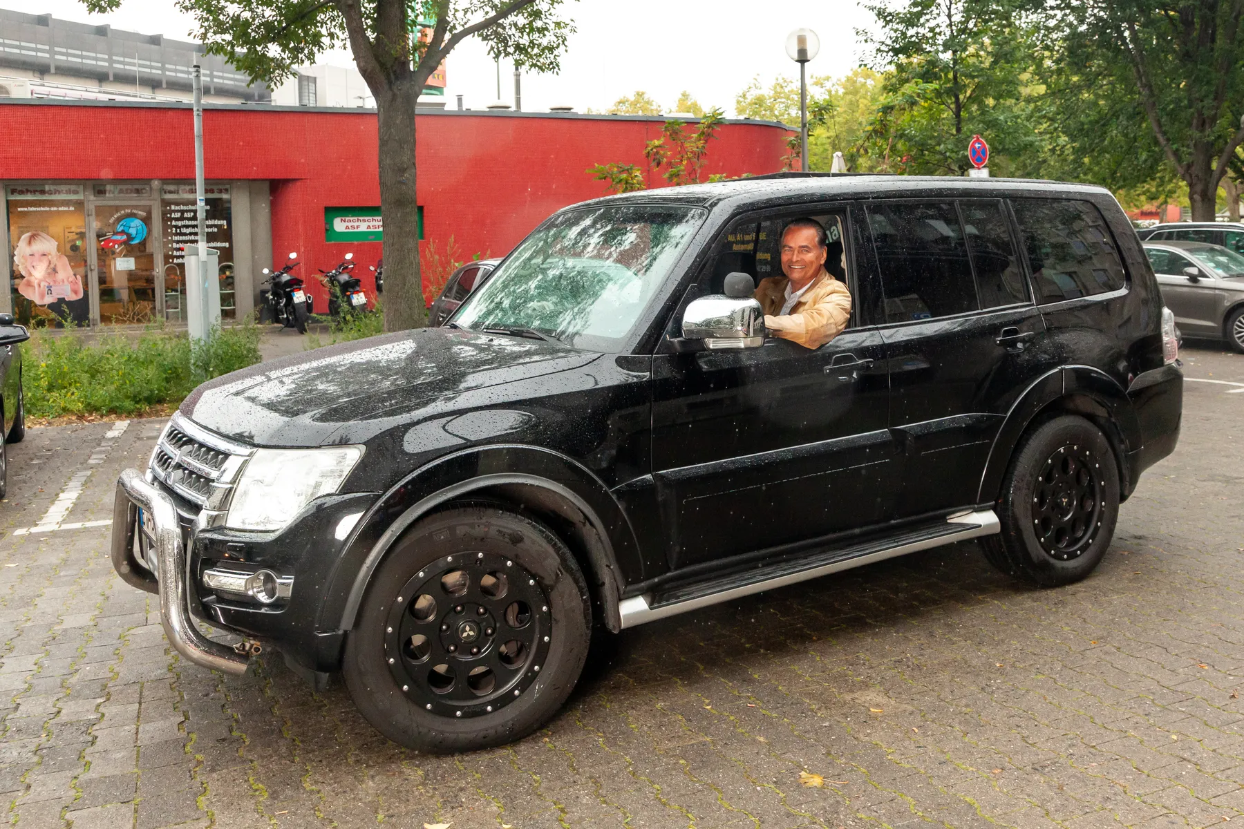 800x800 Ein lächelnder Mann, der auf dem Fahrersitz eines schwarzen Geländewagens sitzt, der auf einem gepflasterten Grundstück mit Bäumen und einem roten Gebäude im Hintergrund geparkt ist.