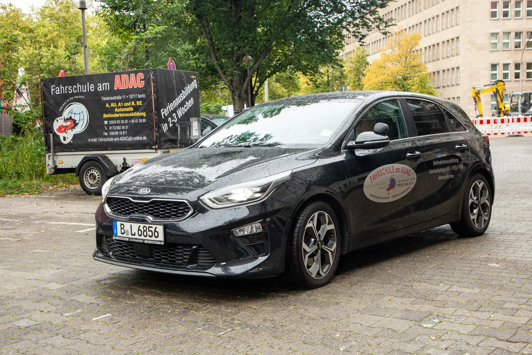 800x800 Ein schwarzer Kia mit Schrägheck und Fahrschullogo ist auf einer nassen Straße geparkt, im Hintergrund sind ein Fahrschulbus und städtische Gebäude zu sehen. Auch Bäume und Bauzäune sind zu sehen.